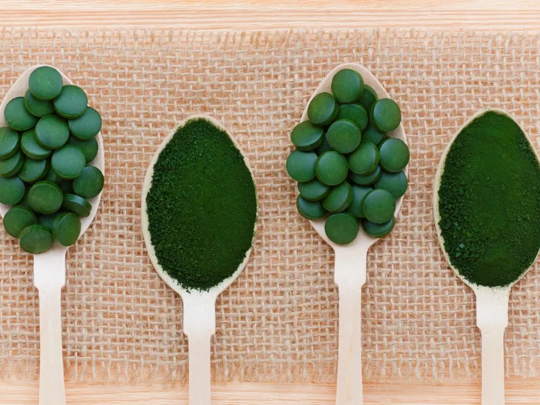 Introbild: Chlorella vs Spirulina Chlorella- und Spirulina-Präparate in Pulver- und Tablettenform auf Holzlöffeln, präsentiert auf einem natürlichen Untergrund.