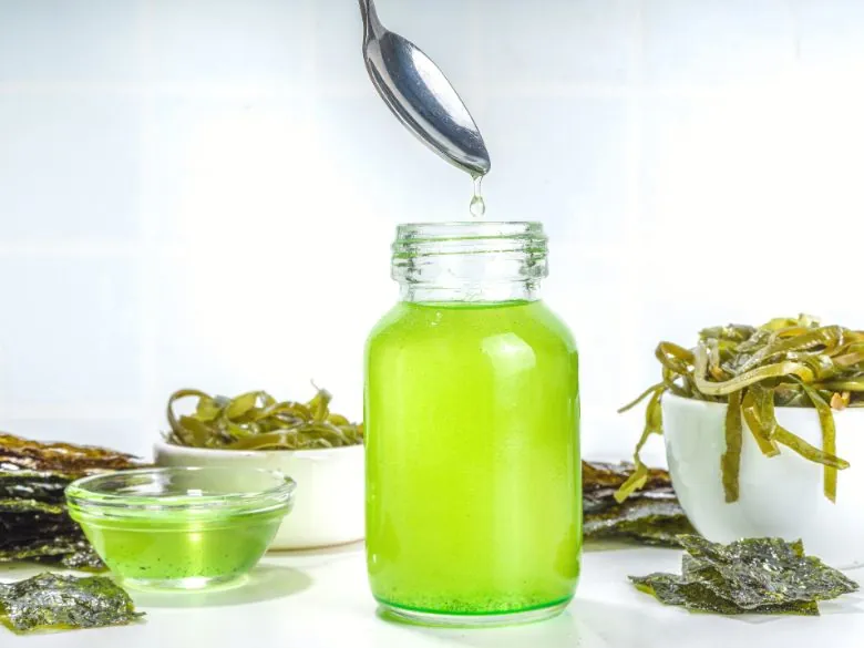 Ein Glas mit grünem Spirulina-Haaröl und Algen im Hintergrund, ideal zur Pflege und Stärkung der Haare bei Haarausfall.